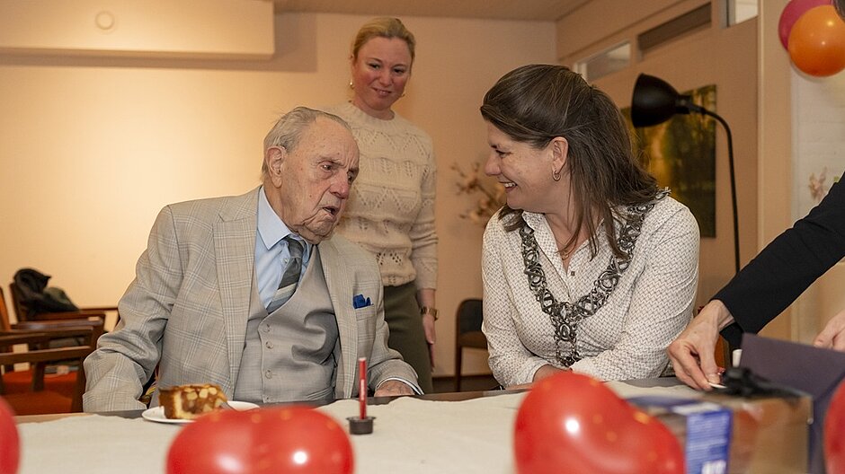 Burgemeester Fleur Gräper op bezoek bij dhr. Tibosch die 100 jaar werd. Op de voorgrond liggen rode hartjes en er staat een stukje appeltaart klaar.