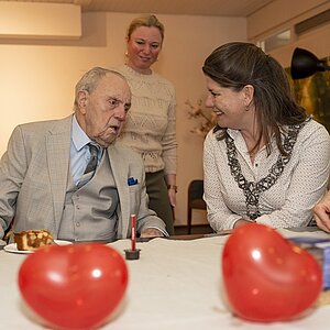 Burgemeester Fleur Gräper op bezoek bij dhr. Tibosch die 100 jaar werd. Op de voorgrond liggen rode hartjes.