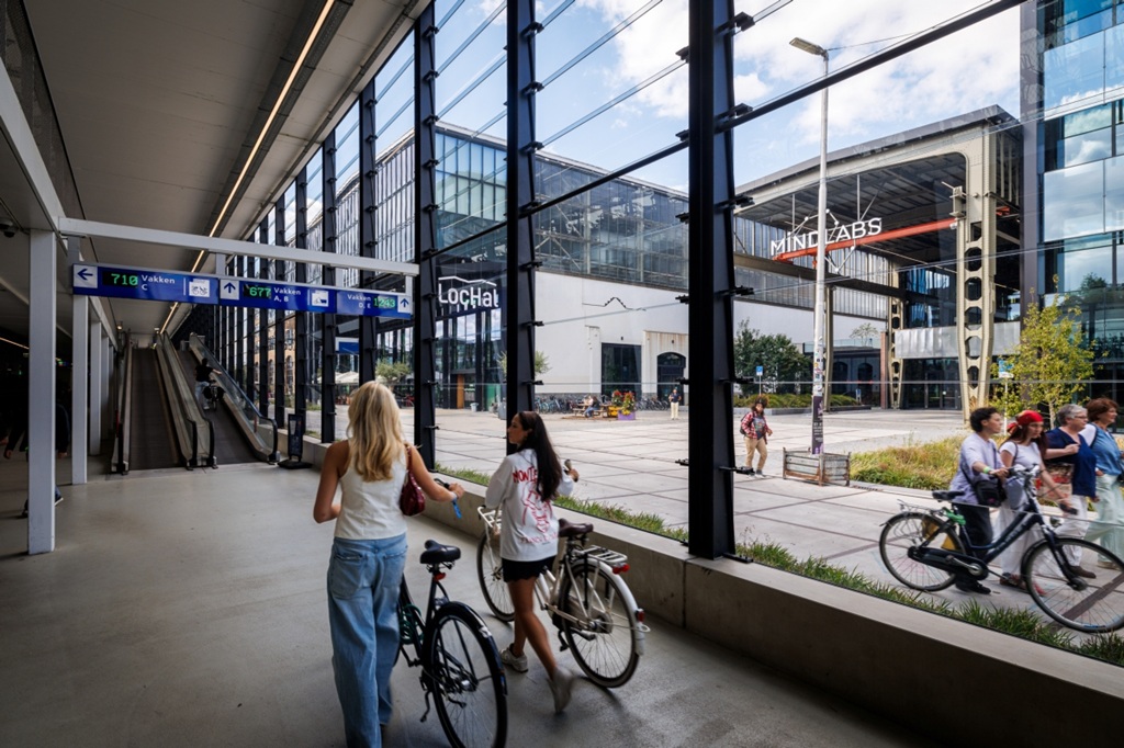 twee dames lopen met hun fiets aan de hand langs grote ramen richting een rolband