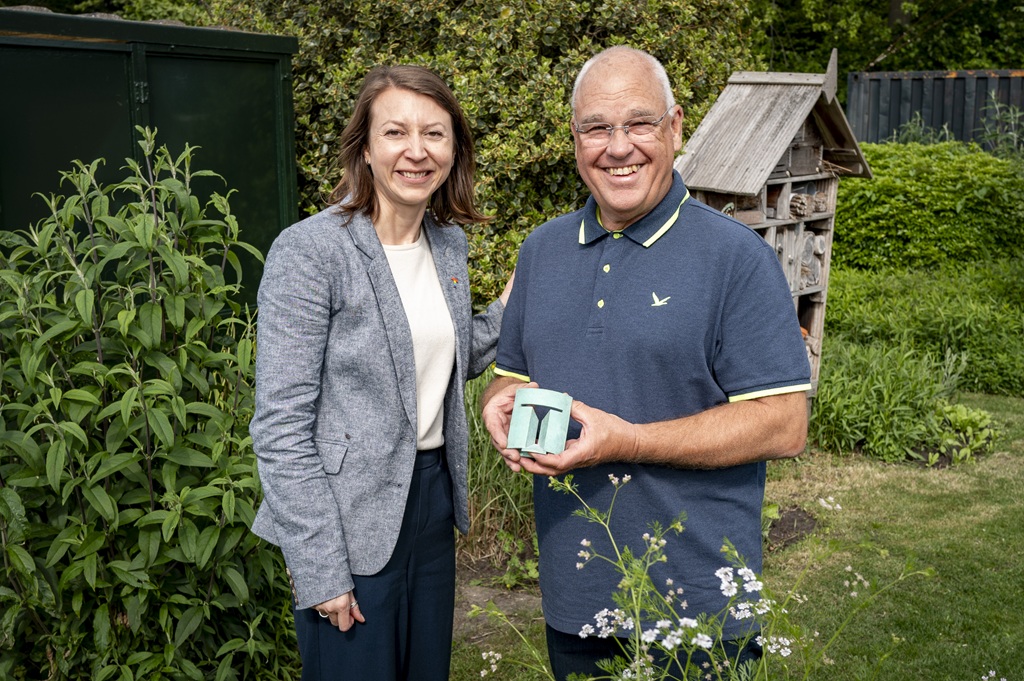 Wethouder Evelien Kostermans en Christ van Geel staan in een tuin, dhr. van Geel houdt de Tilburg Trofee in zijn handen