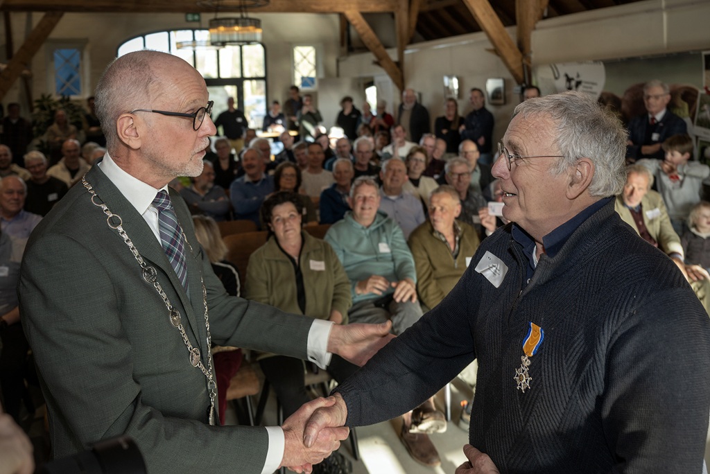 De heer Boink krijgt de Koninklijke onderscheiding uitgereikt in een zaal voor mensen.