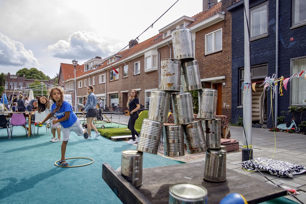 Een jongen met lang haar gooit een stapel blikken om terwijl andere kinderen geamuseerd naar hem kijken.